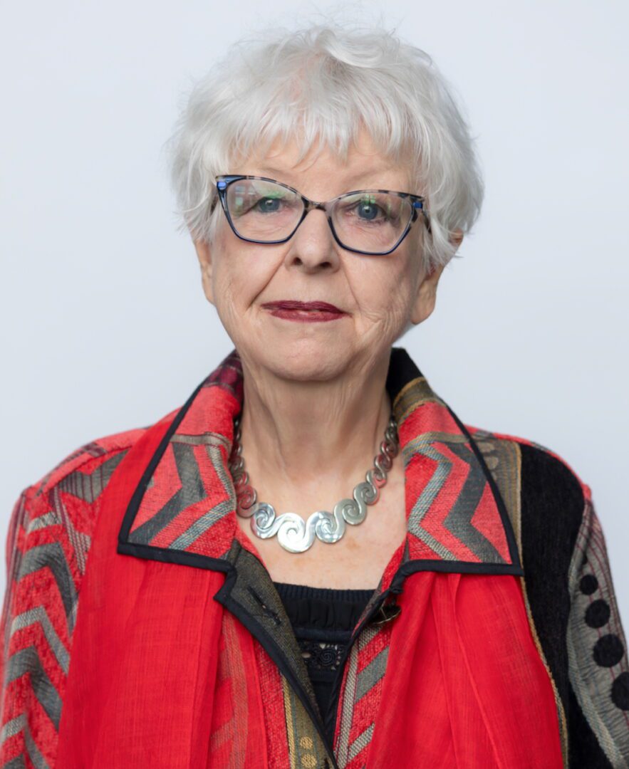 Elderly woman with white hair and glasses wearing a red patterned jacket.